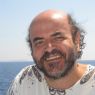 Uśmiechnięty Christos Papadimitriou na tle morza w białej letniej koszuli.